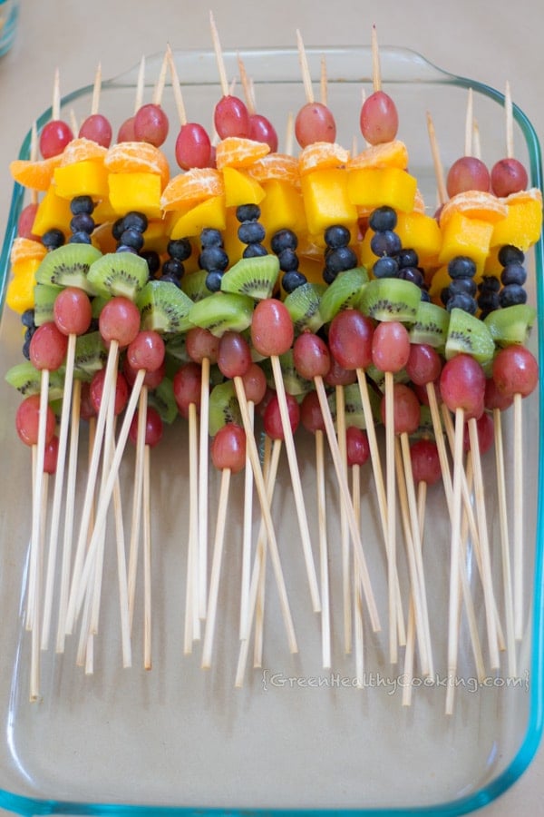 Several fruit kebabs made with grapes, mandarins, mangoes, blueberries and kiwi, arranged in a glass container.