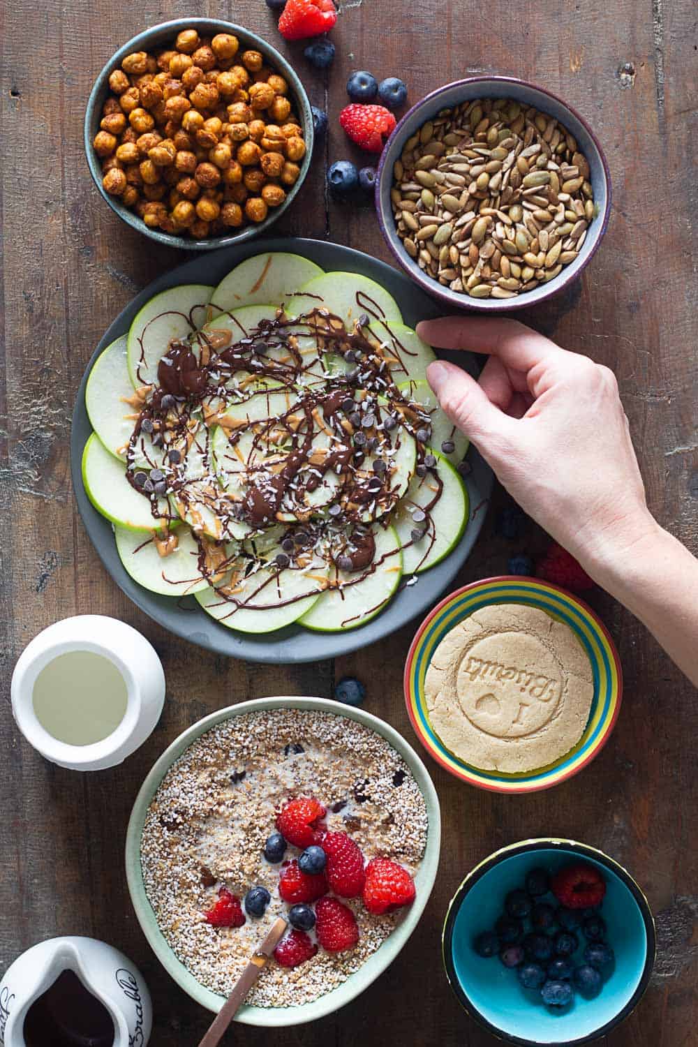 Cereal in a bowl, cookie on a plate, crispy chickpeas in a bowl, seeds in a bowl, all on a wooden table.