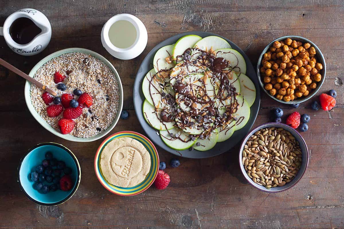 Cereal in a bowl, cookie on a plate, crispy chickpeas in a bowl, seeds in a bowl, all on a wooden table.