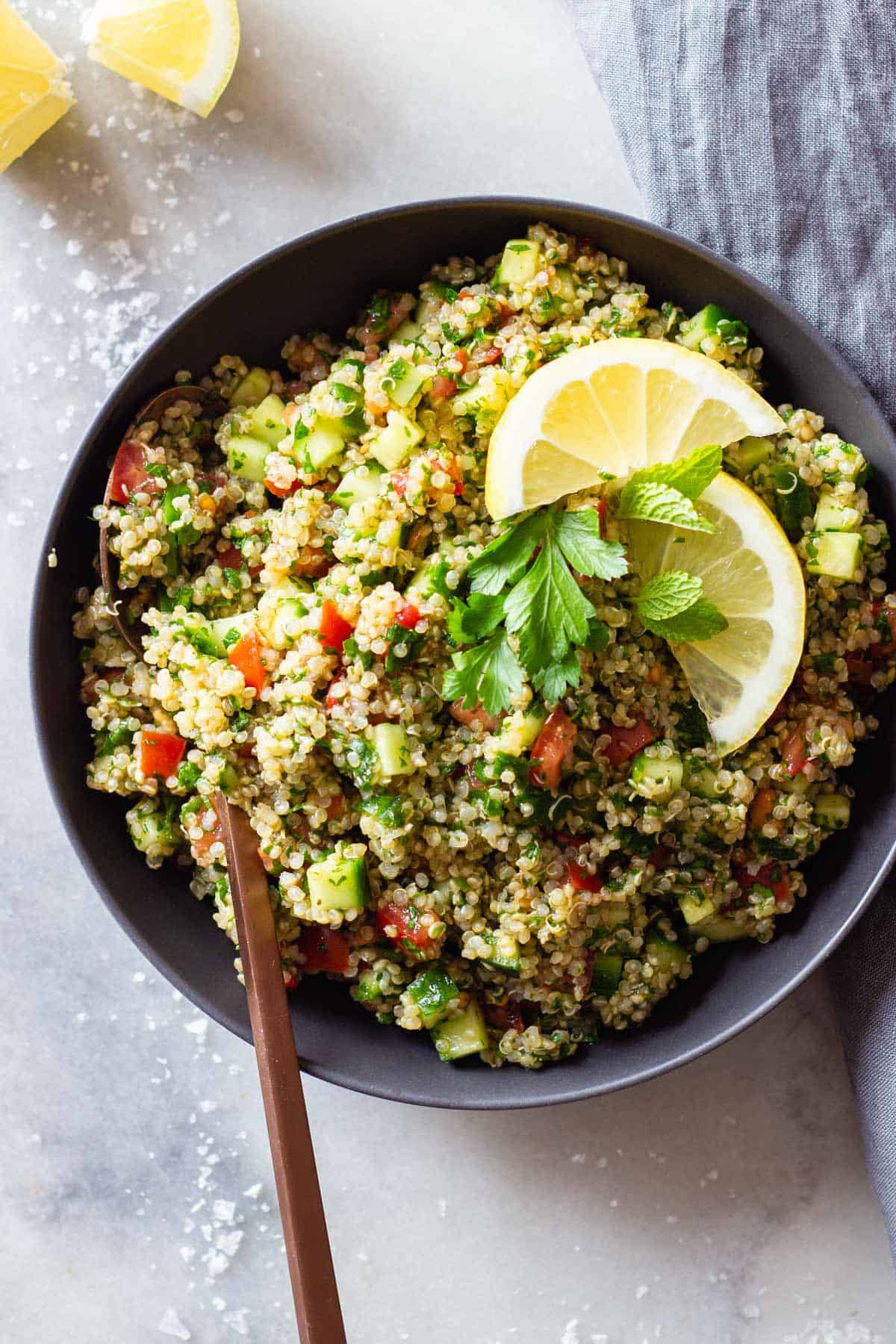 Quinoa Tabbouleh in a gray bowl on a marble surface topped with lemon slices and parsley leaves.