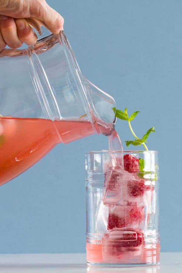 Pink Lemonade poured from a pitcher into a glass filled with ice, raspberries and mint