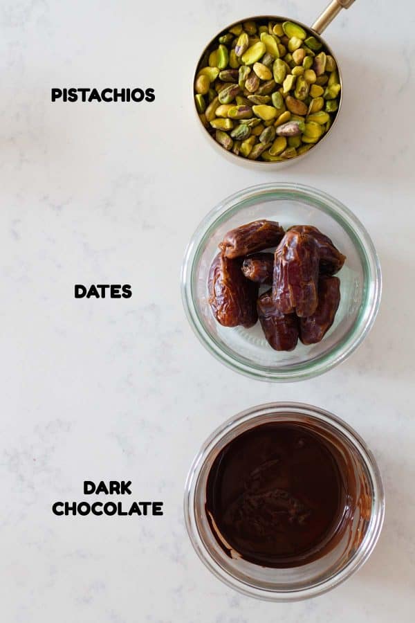 A measuring cup with shelled pistachios, a glass bowl with dates, a glass bowl with melted chocolate and text overlay naming each ingredient.