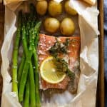 Asparagus, salmon, baby potatoes, a slice of lemon and dill in a ripped parchment paper bag on a baking sheet.