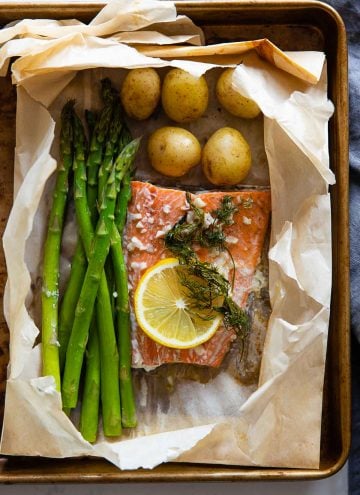 Asparagus, salmon, baby potatoes, a slice of lemon and dill in a ripped parchment paper bag on a baking sheet.