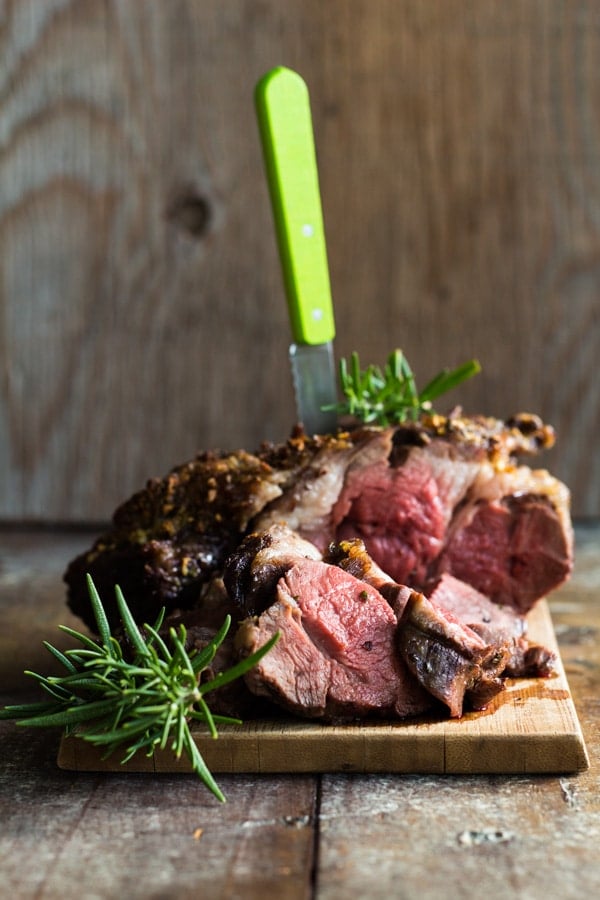 Boneless lamb shoulder roast with rosemary sprigs served on a wood board, and a knife inserted on top. 