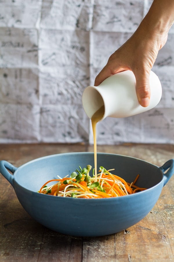 Hand pouring dressing from a white jar over Sunflower Carrot Salad in a blue bowl.