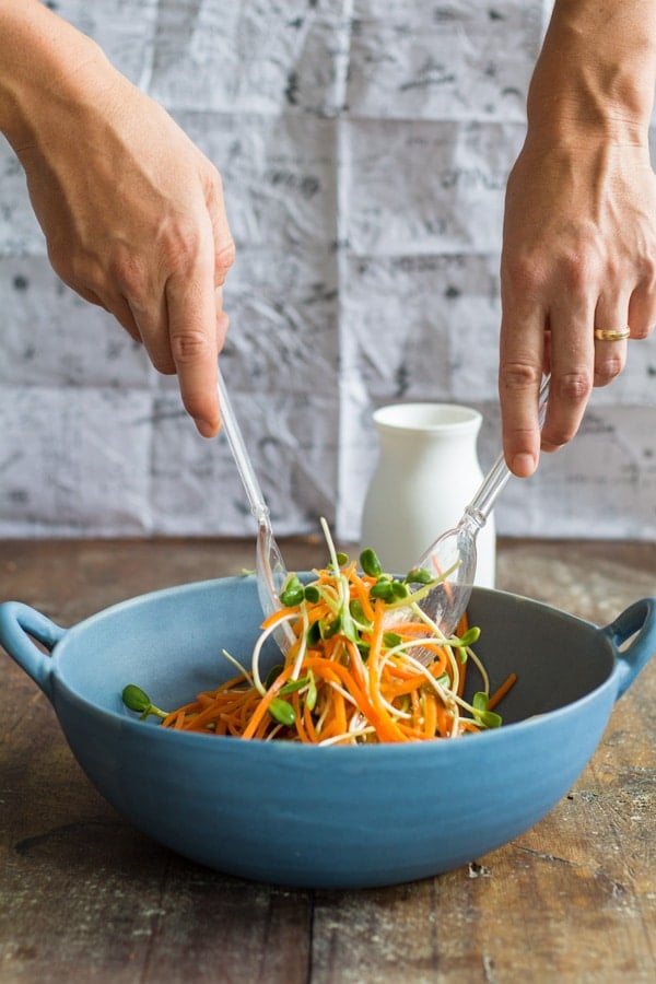 Hands tossing Sunflower Carrot Salad with a salad spoon and fork in a blue bowl.
