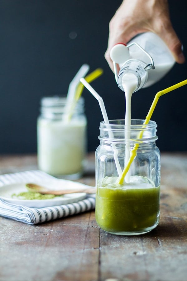 Hand pouring milk in an Iced Maple Matcha Latte glass jar with two straws.