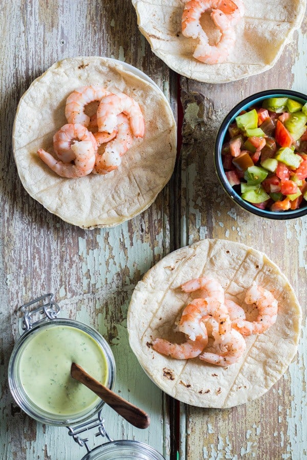 Shrimp Tacos with a small cup of Cilantro Lime Salsa and a cup of chopped tomatoes on a rustic wooden board.