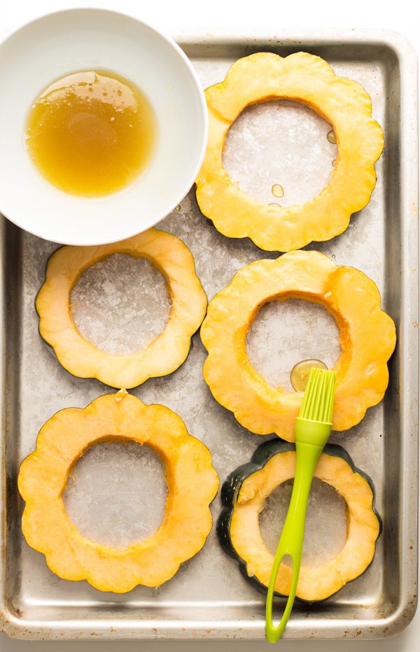 Acorn squash slices on a baking sheet, melted butter in a bowl and a basting brush.