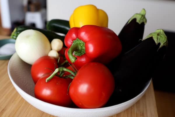 Tomatoes, bell peppers, eggplants, zucchinis, onion, garlic in a white bowl.