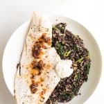 Top view of Haddock with Black Quinoa Risotto and creme fraiche on a white plate, and a cup with creme fraiche with a wooden spoon.