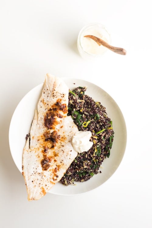 Top view of Haddock with Black Quinoa Risotto and creme fraiche on a white plate, and a cup with creme fraiche with a wooden spoon.