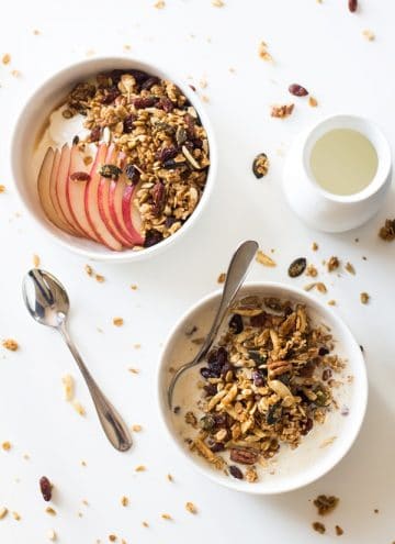 Healthy Homemade Granola in a bowl with milk and a spoon, and topping another bowl of yogurt and sliced apple. A spoon and milk jar next to it.