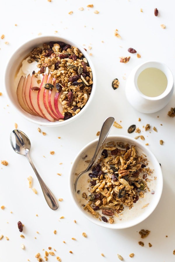 Healthy Homemade Granola in a bowl with milk and a spoon, and topping another bowl of yogurt and sliced apple. A spoon and milk jar next to it.