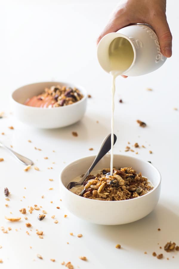 Hand pouring milk from a white jar on a bowl of Homemade Granola with a spoon. Another bowl of granola in the background.