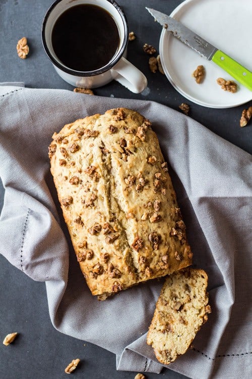 Top view of sliced Banana Bread with Maple Candied Walnuts, a cup of coffee, and an empty plate with a knife.