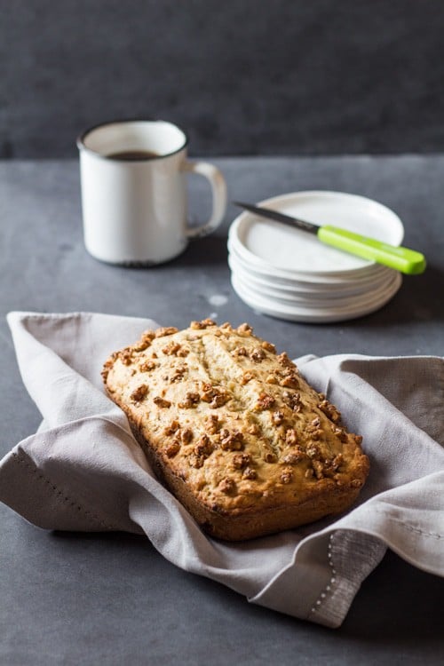 Freshly baked Banana Bread with Maple Candied Walnuts on a beige napkin, a cup of coffee, and some serving plates with a knife.