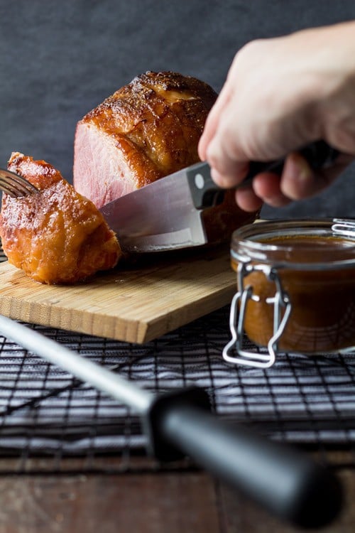 Hand cutting a freshly baked ham with a knife on a cutting board, and an open jar with gravy.