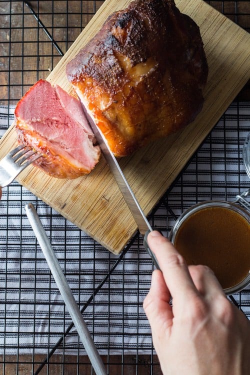 Top view of hand cutting freshly baked ham on a cutting board, and an open jar of gravy.