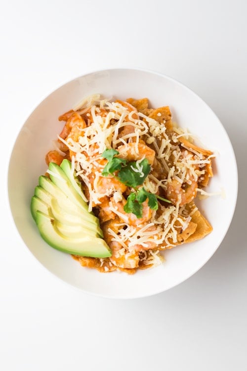 A bowl of Mexican Breakfast Chilaquiles con Camarones with shredded cheese and avocado slices.