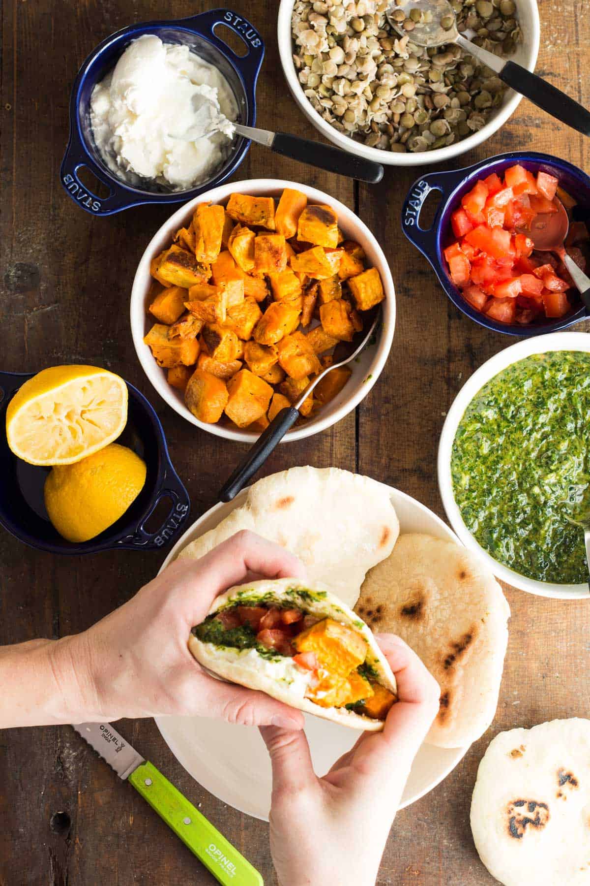 Hands holding a Pita With Sweet Potato Lentil Filling and Out of This World Tahini Herb Sauce, and all the ingredients in bowls.