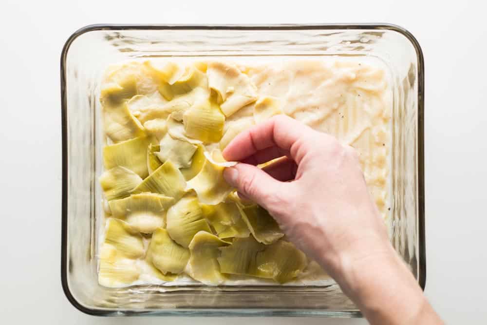 Hand putting artichoke pieces over lasagna sheets in a baking dish.