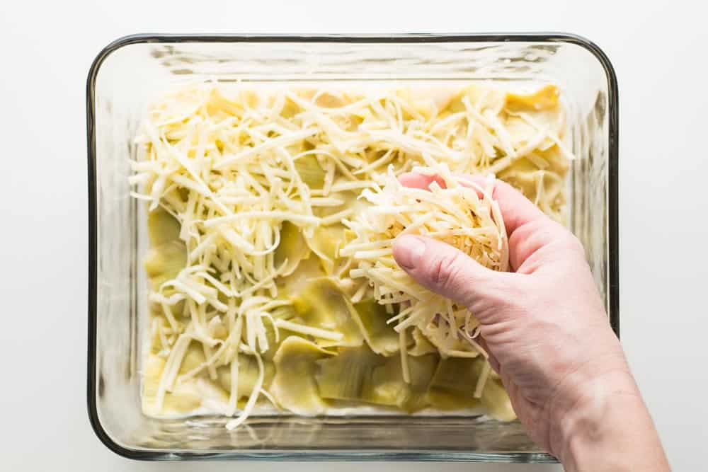 Hand putting shredded cheese over layer of artichoke in a baking dish.