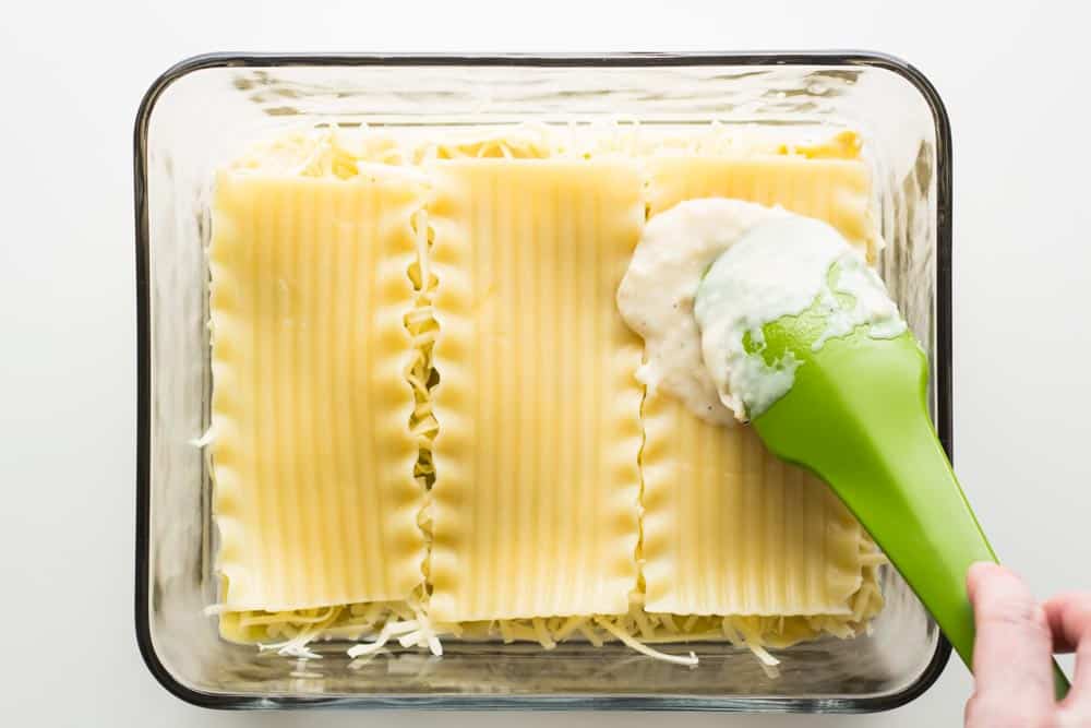 Spatula spreading bechamel sauce on top of lasagna sheets in a baking dish.