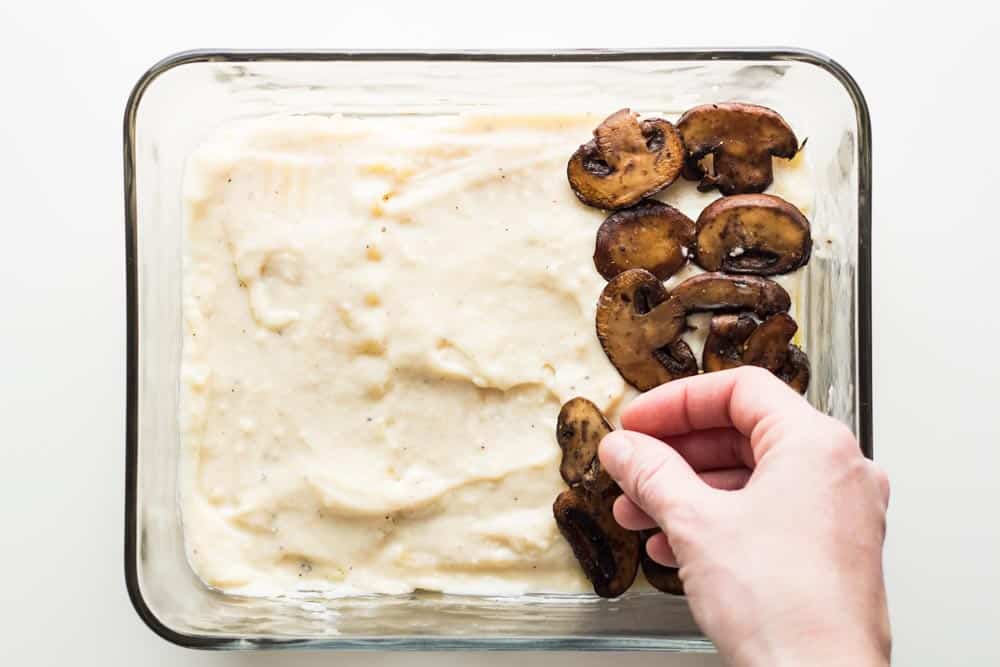 Hand placing mushrooms on top of bechamel sauce layer of lasagna in a baking dish.
