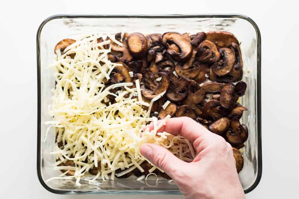 Hand spreading shredded cheese on top of mushrooms layer of lasagna in a baking dish.
