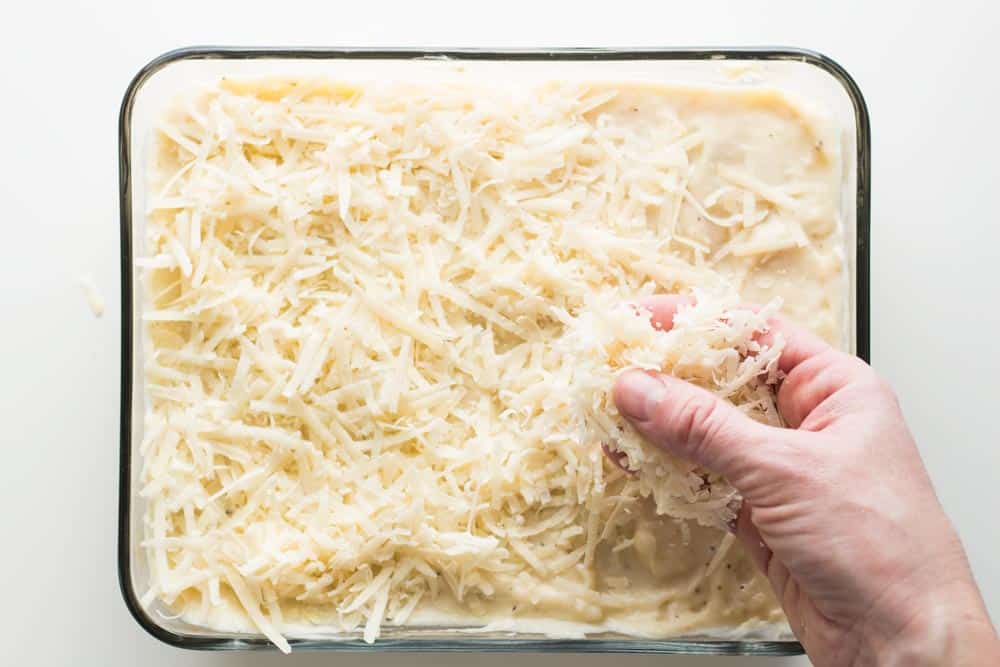 Hand spreading shredded cheese on top of bechamel sauce layer of lasagna in a baking dish.