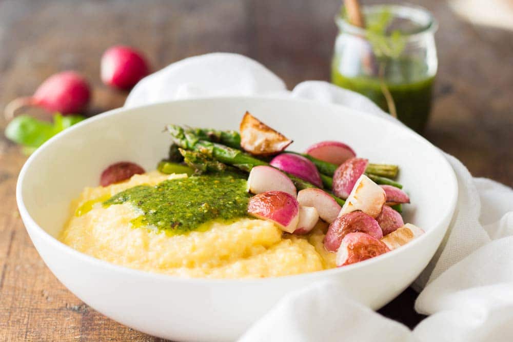 Creamy Parmesan Polenta with Grilled Vegetables and 5-Minute Basil Pistachio Pesto in a white bowl, and an open jar of pesto sauce in the background.