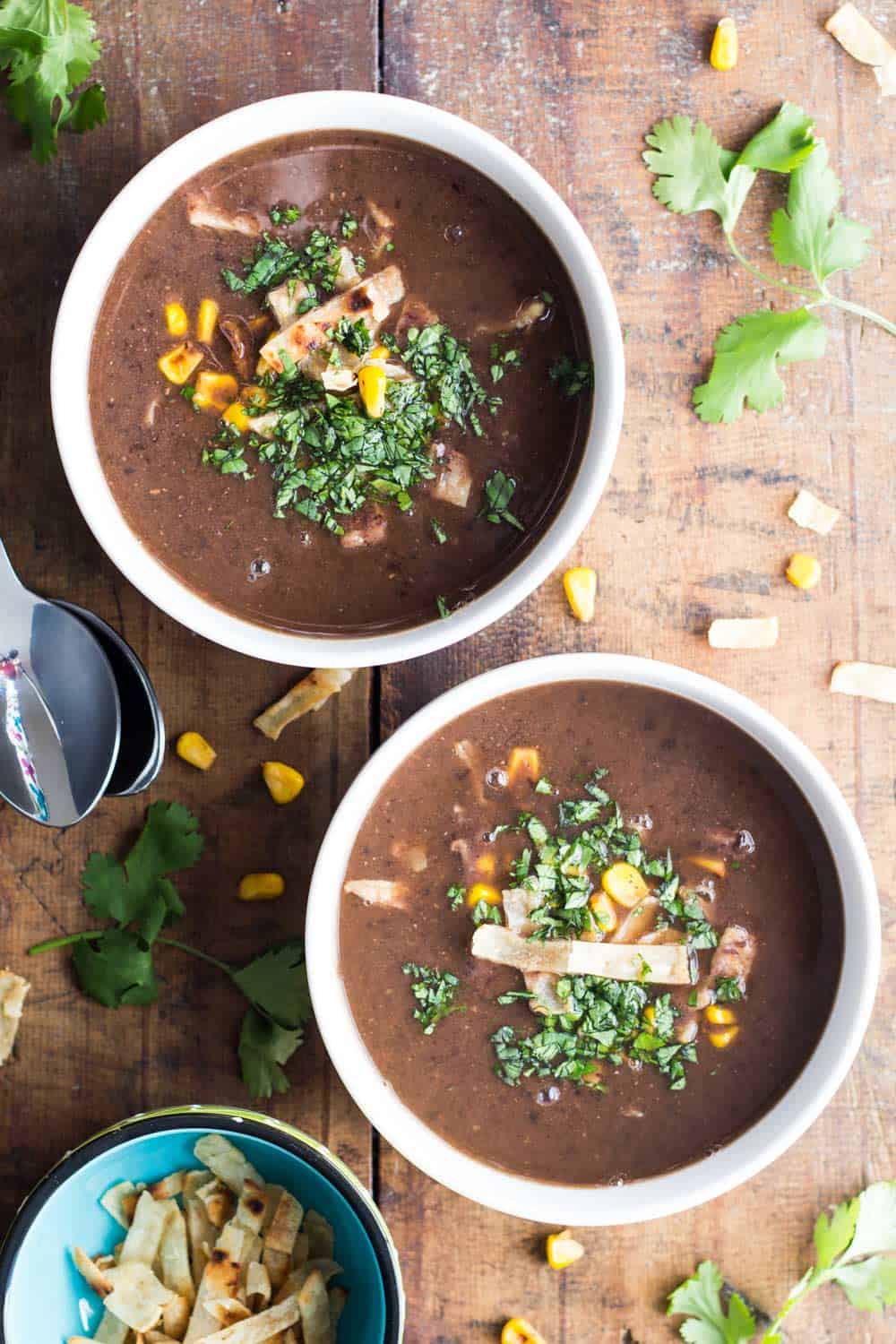 Two bowls of Mexican Black Bean Soup on a wooden board, two spoons and a small bowl of tortilla strips.