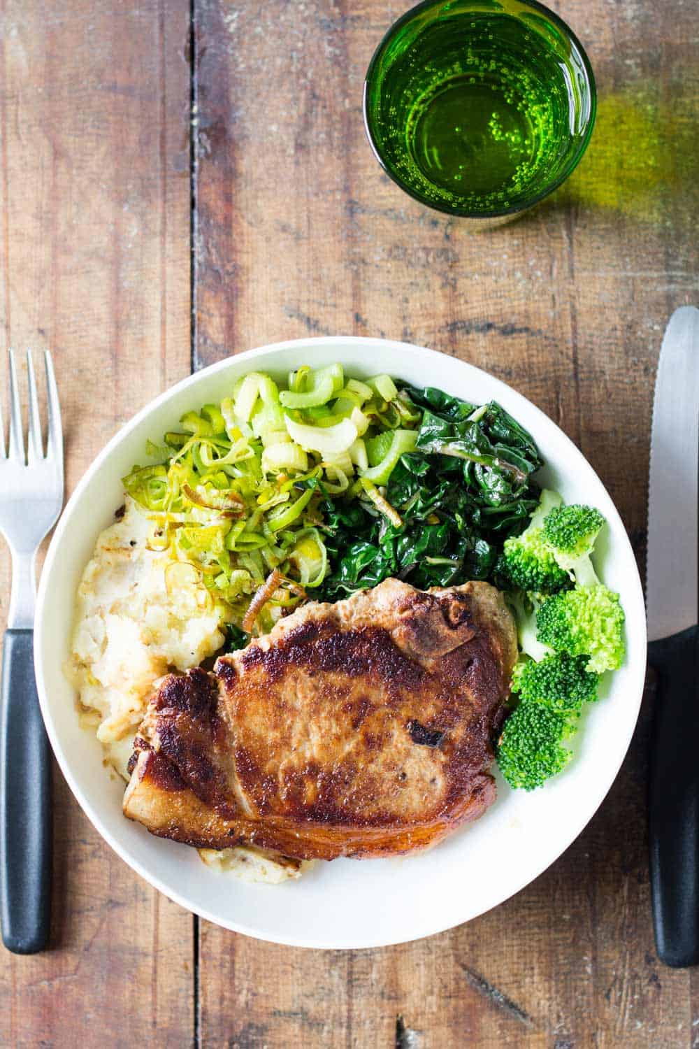 Perfect Pork Chop with mashed potatoes, celery, chard and broccoli, on a wooden table with a fork and knife, and a glass of water.