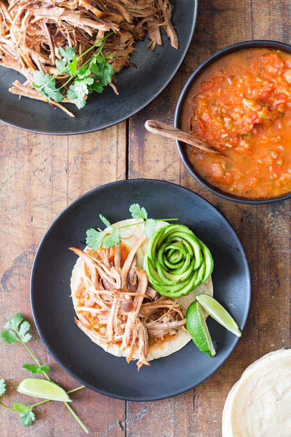 Top view of a pulled pork taco, a bowl of red salsa with a wooden spoon, and part of a plate with pulled pork.