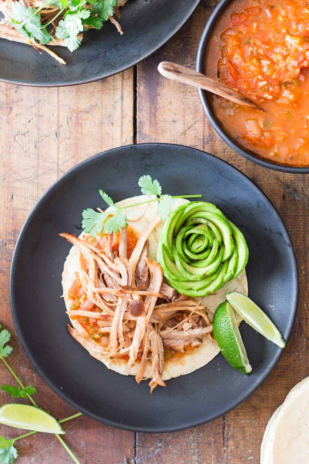 A pulled pork taco with salsa and flower-shaped avocado slices, and slice lime on a black plate. Part of red salsa bowl with a wooden spoon.