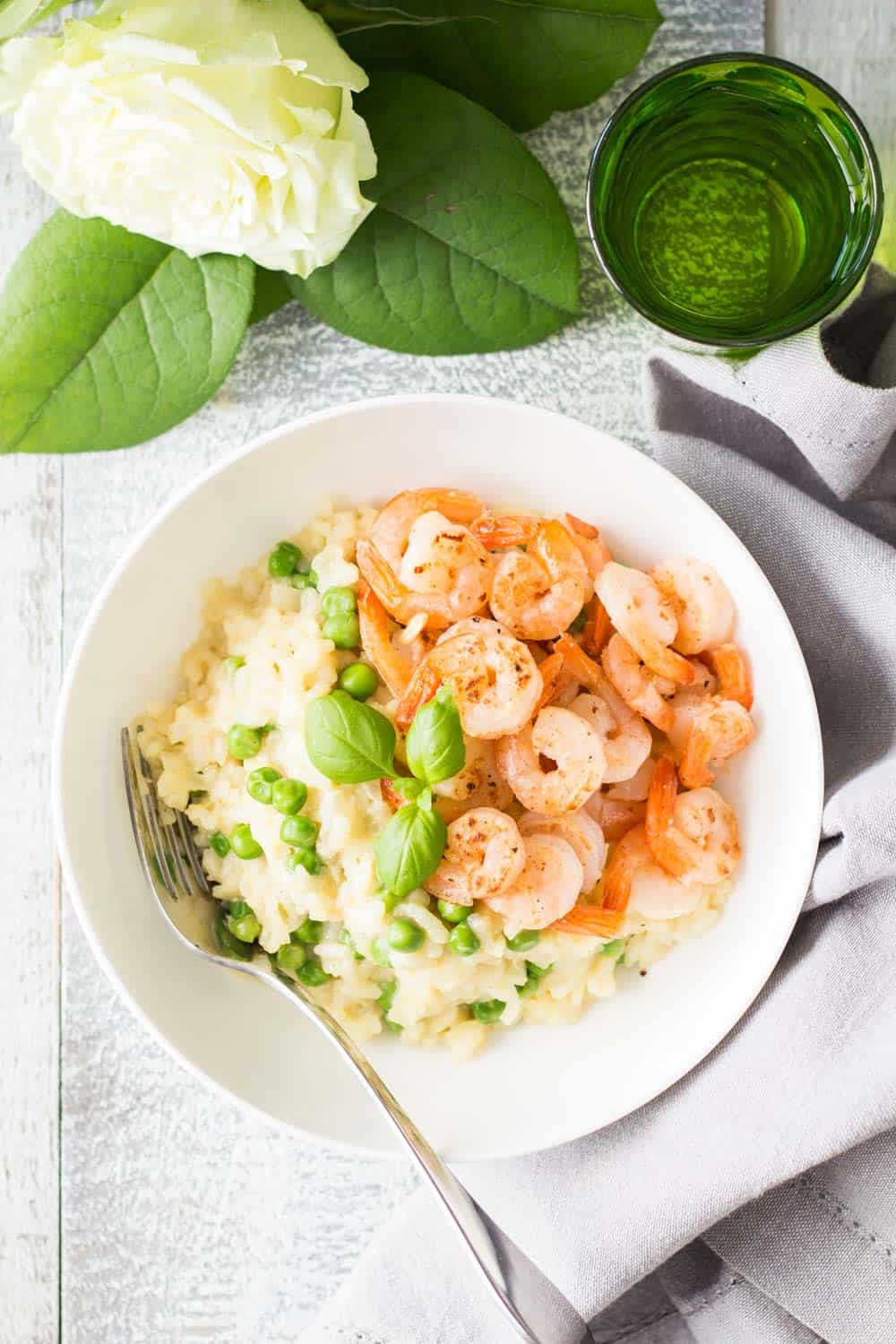 Top view of Shrimp and Pea Risotto on a white plate with a fork, a glass of water and a flower.