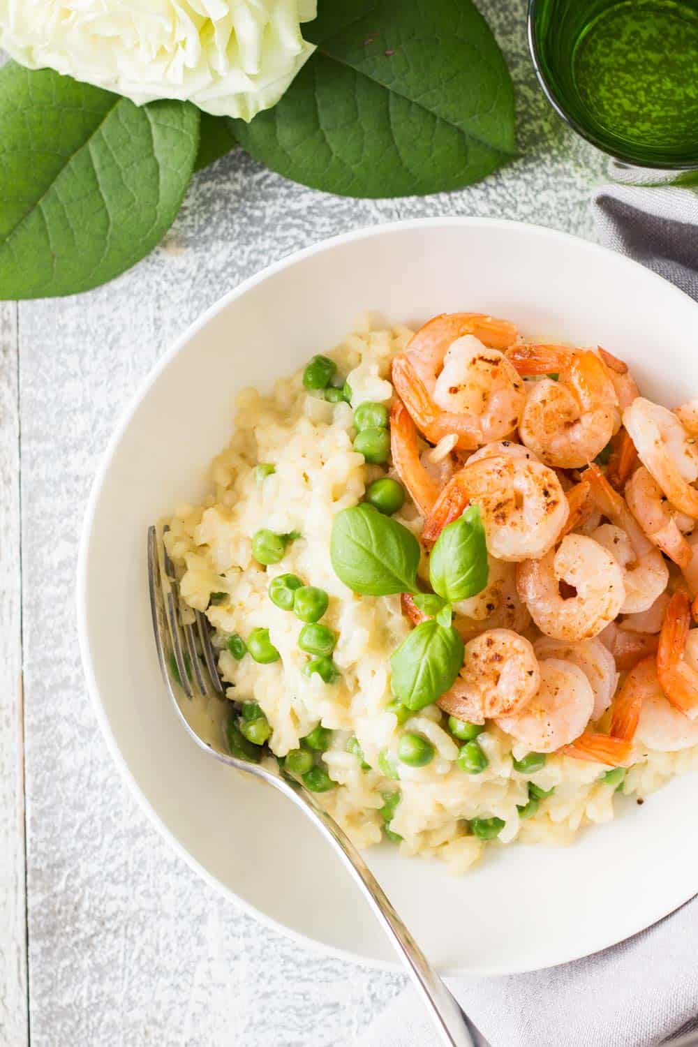 Top view of Shrimp and Pea Risotto on a white plate with a fork, a glass of water and a flower.