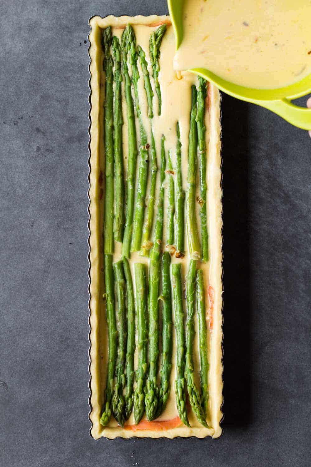 pouring egg batter into a tart pan over asparagus spears to make quiche
