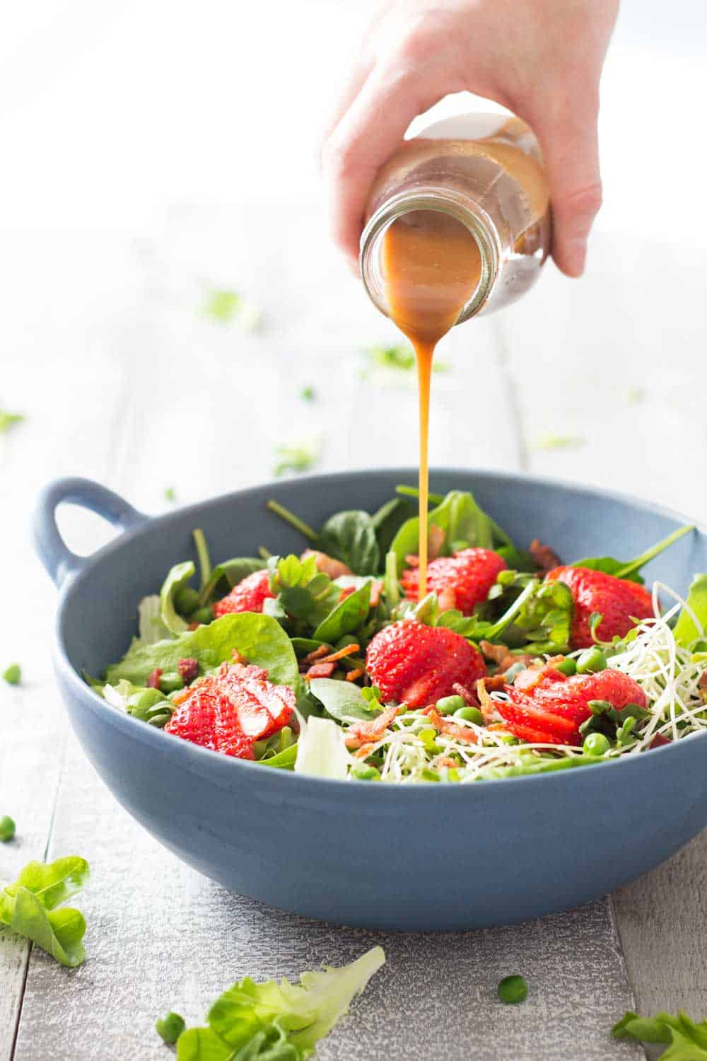 Hand pouring Maple Mustard Balsamic Dressing over Strawberry Bacon Spring Salad in a blue bowl.