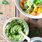 Top view of jar of Cilantro Pesto with a wooden spoon, and part of a bowl of rainbow shell pasta topped with Cilantro Pesto.