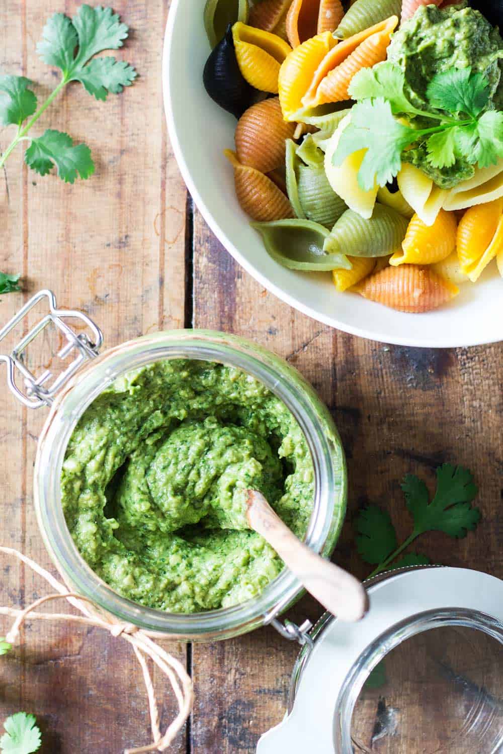 Top view of jar of Cilantro Pesto with a wooden spoon, and part of a bowl of rainbow shell pasta topped with Cilantro Pesto.