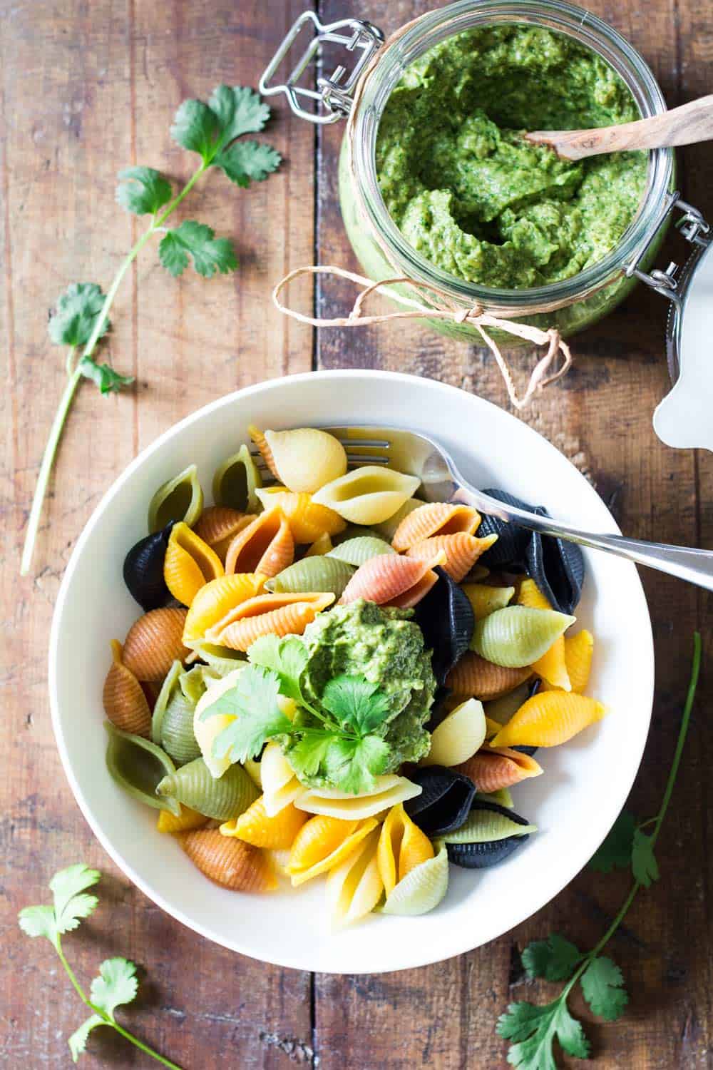 Top view of bowl of rainbow shell pasta topped with Cilantro Pesto, and a jar of Cilantro Pesto with a wooden spoon.