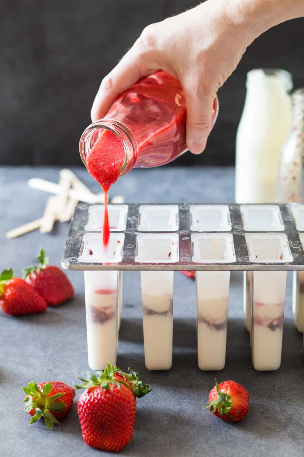 Hand pouring strawberry mix into popsicle molds containing greek yogurt and blueberry mix.