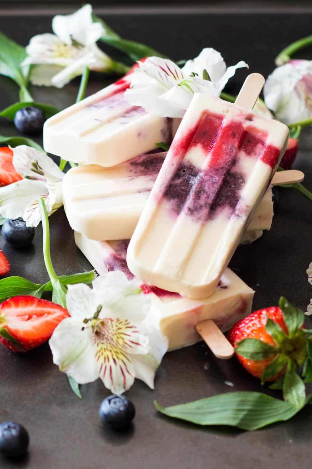 Close up of Greek Yogurt Berry Popsicles laid in a pile with fresh flowers, strawberries and blueberries.