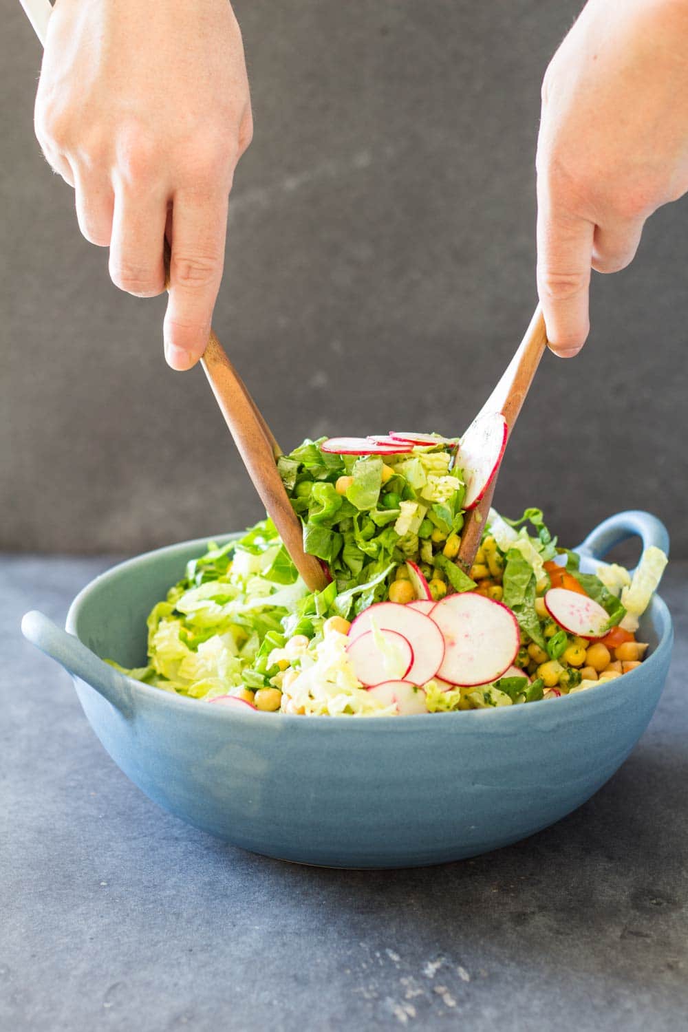 Hands tossing Romaine Lettuce Salad With Herb Vinaigrette with a wooden salad fork and spoon in a blue ceramic bowl.