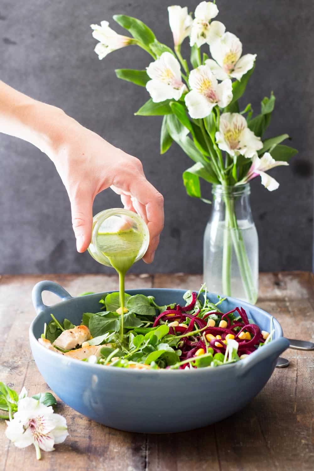 Hand pouring Basil Clementine Dressing over Watercress Salad in a blue bowl, and a vase with flowers.