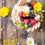 Top view of Acai Bowl topped with fresh fruit and flowers, on a rustic wood table with flowers, and text overlay.