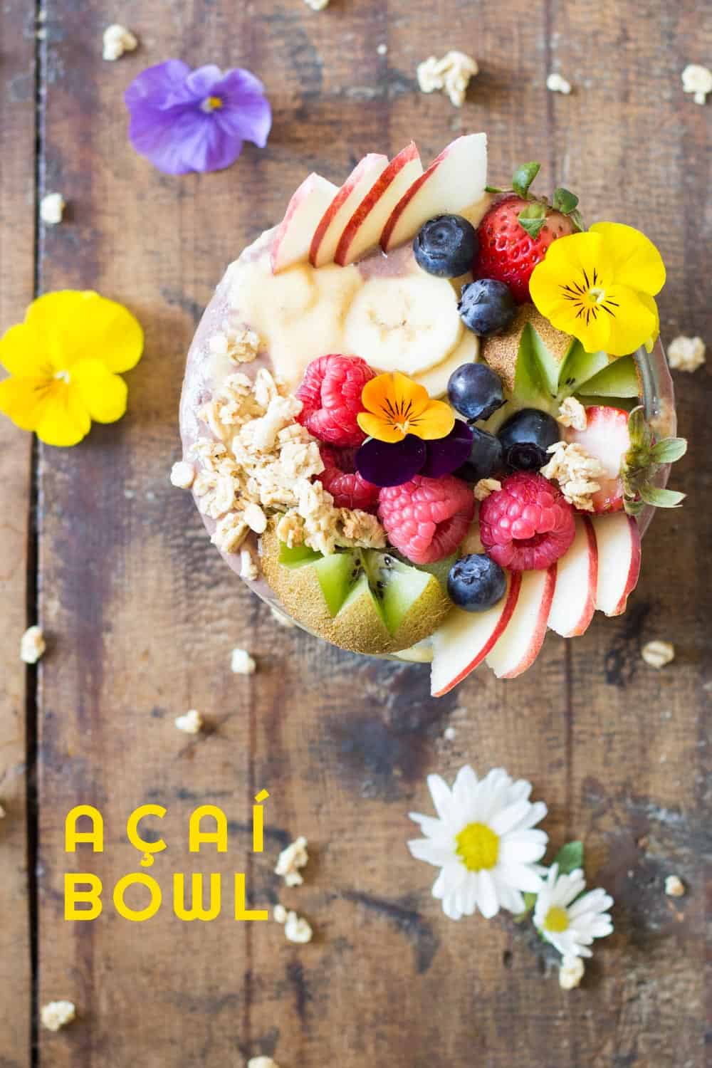 Top view of Acai Bowl topped with fresh fruit and flowers, on a rustic wood table with flowers, and text overlay.